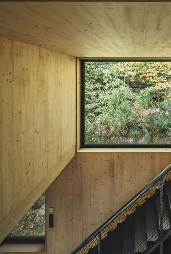Interior staircase detail with black metal mesh railing against light spruce wood panels and a window showing forest foliage.