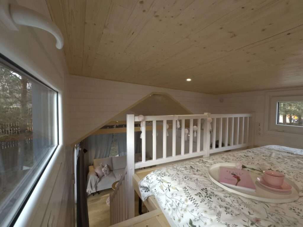 Comfortable sleeping loft in the Smile house with a low wooden ceiling and a view overlooking the living area.