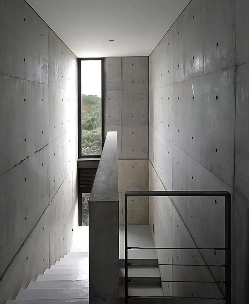 Interior view of the narrow fair-faced concrete staircase inside the 4x4 House, with signature tie-rod holes and a slender vertical window letting in natural light.