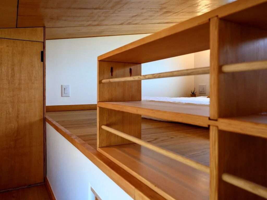 Wooden storage shelving unit acting as a protective guardrail on the edge of a tiny home loft mezzanine.