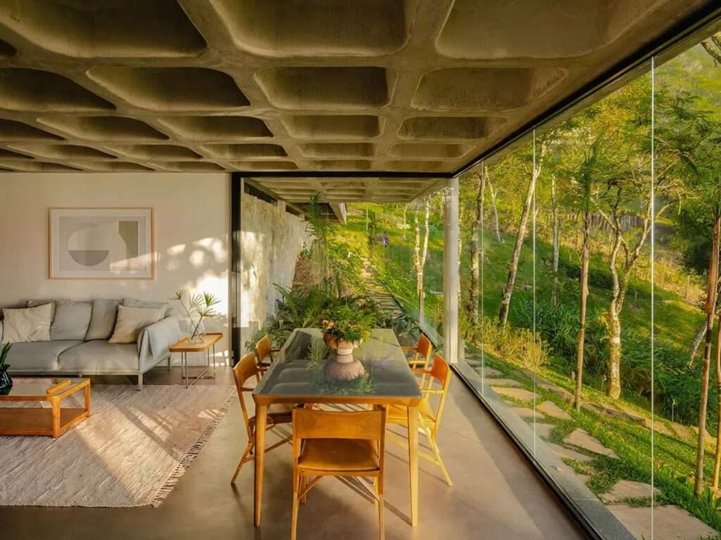 View from a lower dining area looking out through glass walls at a stone wall and tropical plants, under a concrete waffle slab ceiling.