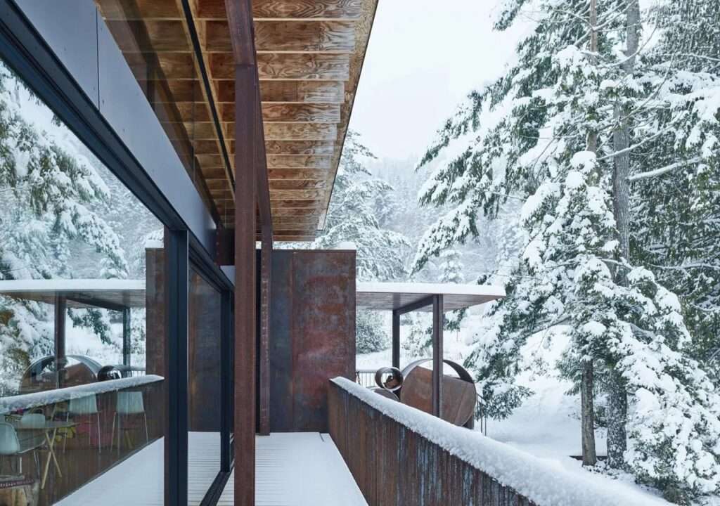Perspective view from the Daisy House balcony showing the weathered steel railing and snow-laden evergreen trees.