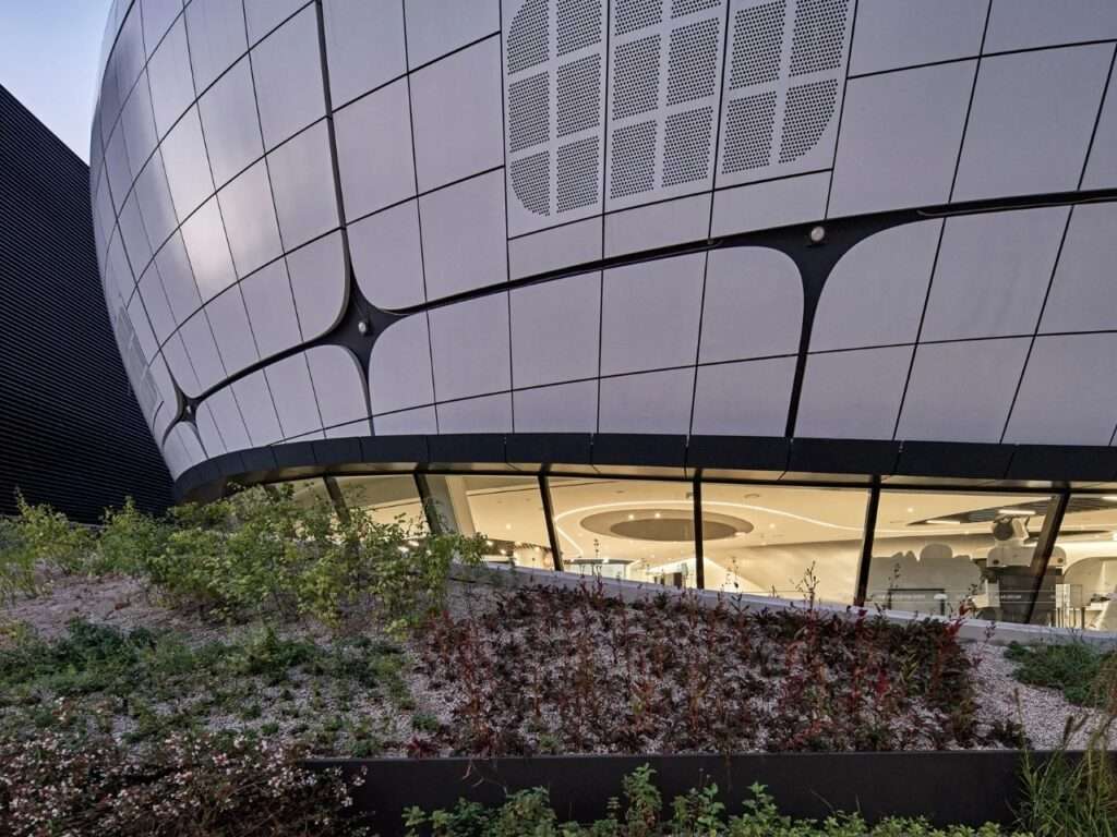 Detailed architectural view of the double-curved metal panels meeting the structural steel grid connections above a curved glass window band at ground level.