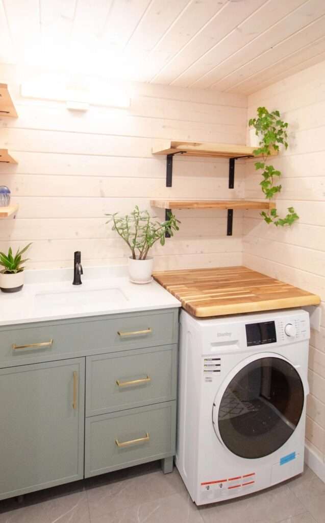 Modern tiny house bathroom featuring a sage green vanity with gold handles, a white undermount sink, floating raw wood shelves, and a compact front-load washing machine.