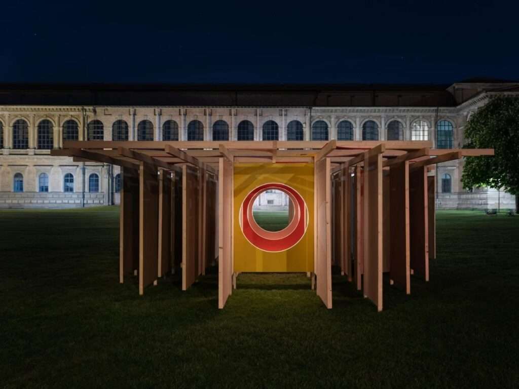 Night photograph of the DELULU installation under a dark sky, with warm artificial lighting illuminating the central circular tunnel through the timber and jute maze, set against the glowing windows of the Alte Pinakothek.