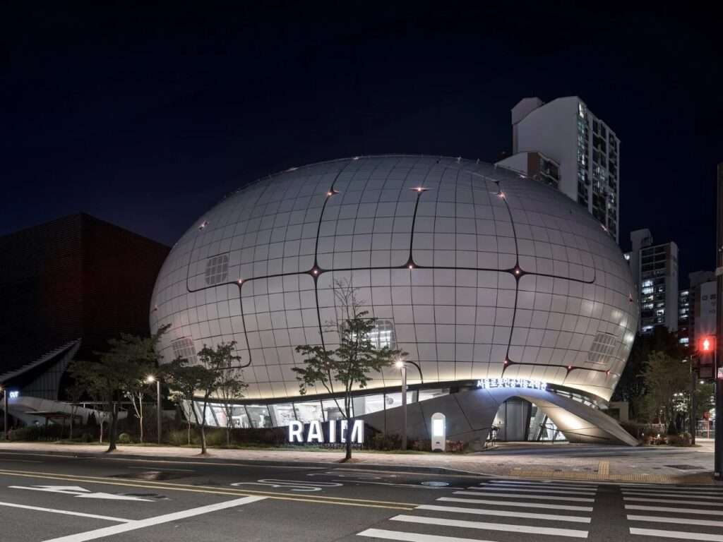 Nighttime exterior photograph of the illuminated spherical Seoul Robot and AI Museum with the glowing "RAIM" sign in front.