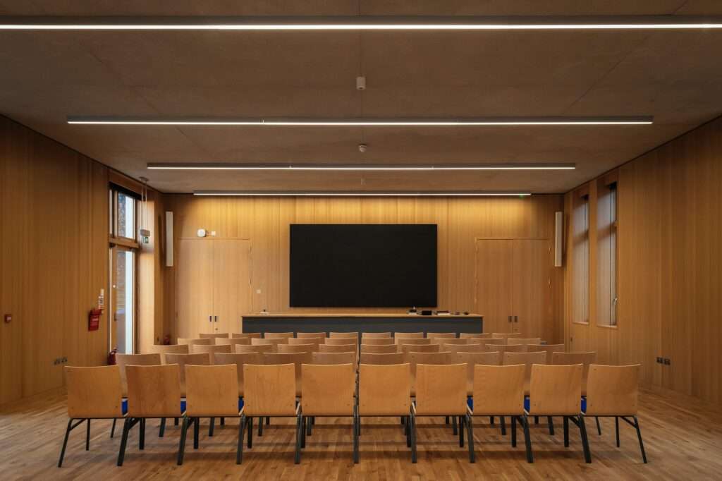 A symmetrical interior view of a modern 120-seat lecture theatre with timber paneled walls, a large digital screen, and rows of wooden chairs.