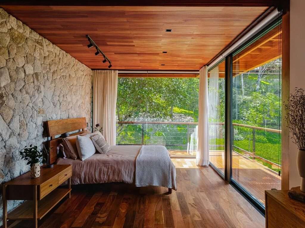 Master bedroom interior with a rustic stone wall, wooden floors, and a balcony door revealing the dense forest.