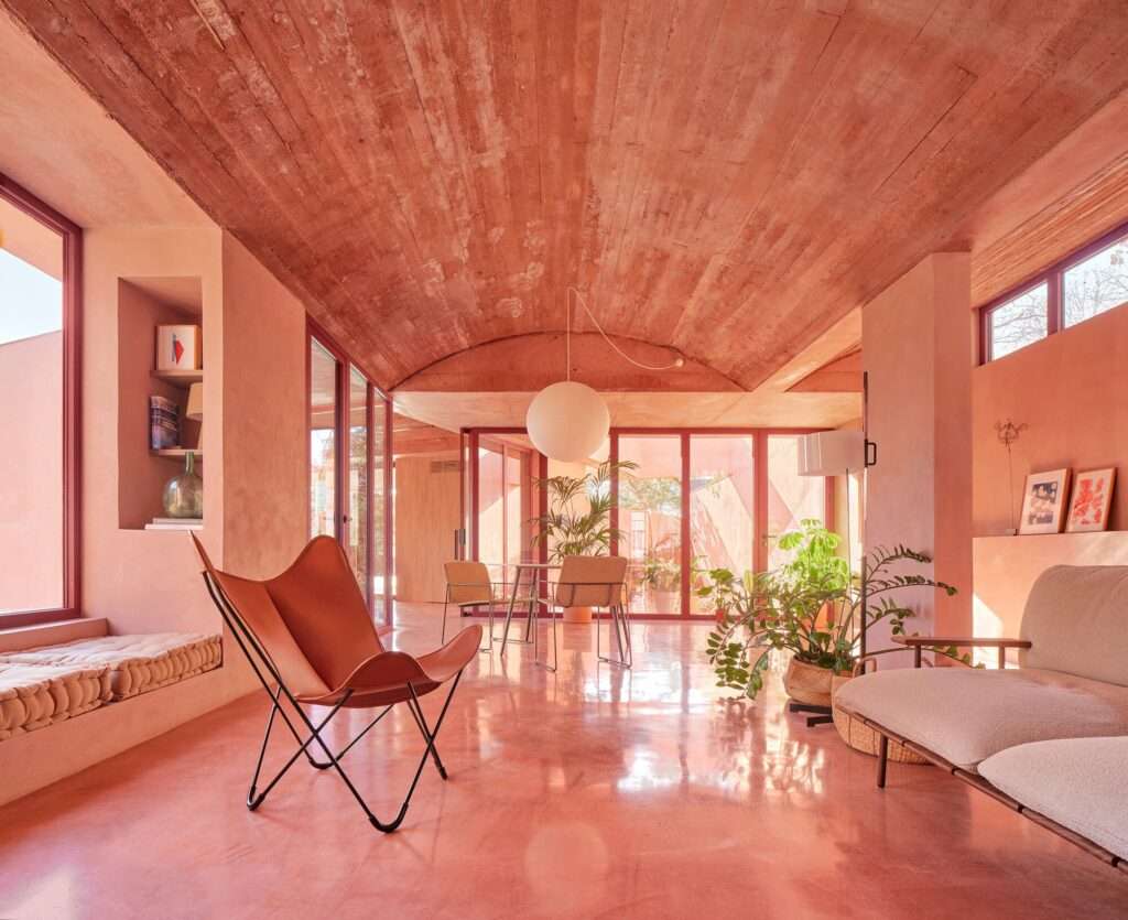 Spacious open-plan living room with red concrete vaulted ceiling, large glass doors opening to a courtyard, minimalist furniture, and a classic butterfly chair.