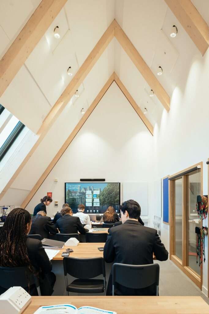 Bright modern school classroom with an steeply angled high ceiling supported by light wood glulam beams and students sitting at desks facing an interactive screen.