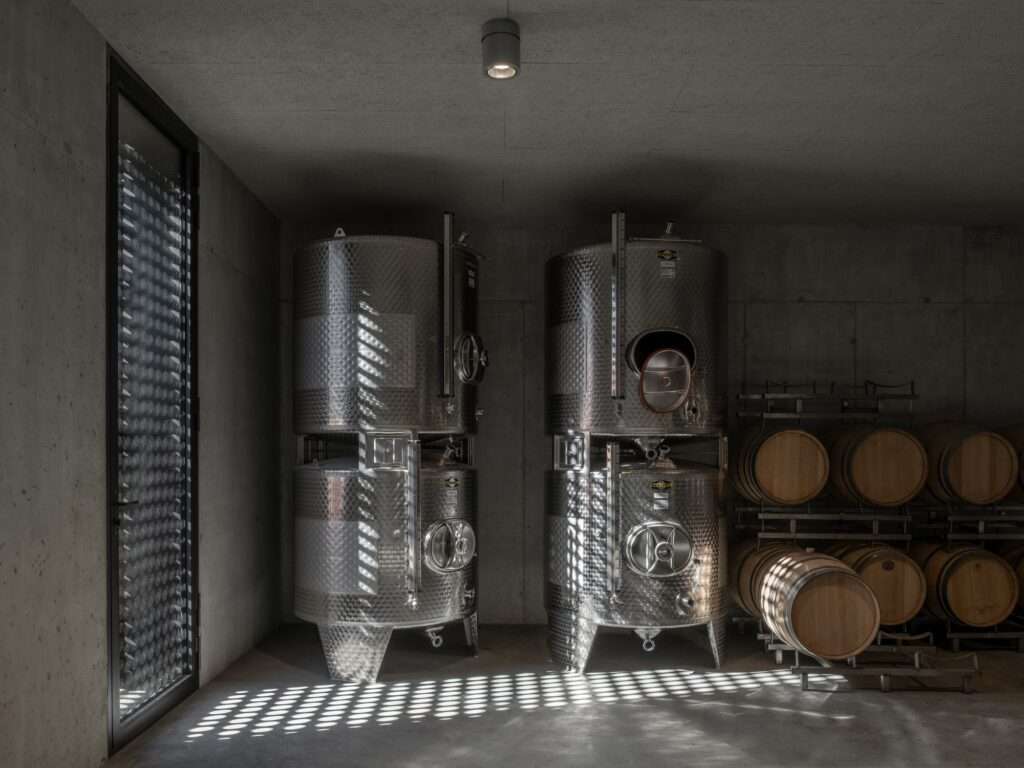 A view of the storage area at Cantina Hauser featuring stacked stainless steel wine tanks and oak barrels, illuminated by natural light through the perforated facade.
