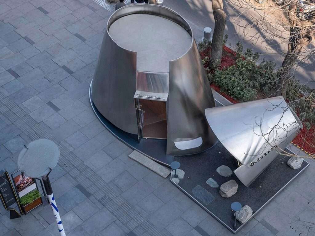 High-angle top-down view of the Kurasu coffee pavilion, showing the asymmetric cone shapes and its layout on the plaza gravel bed.