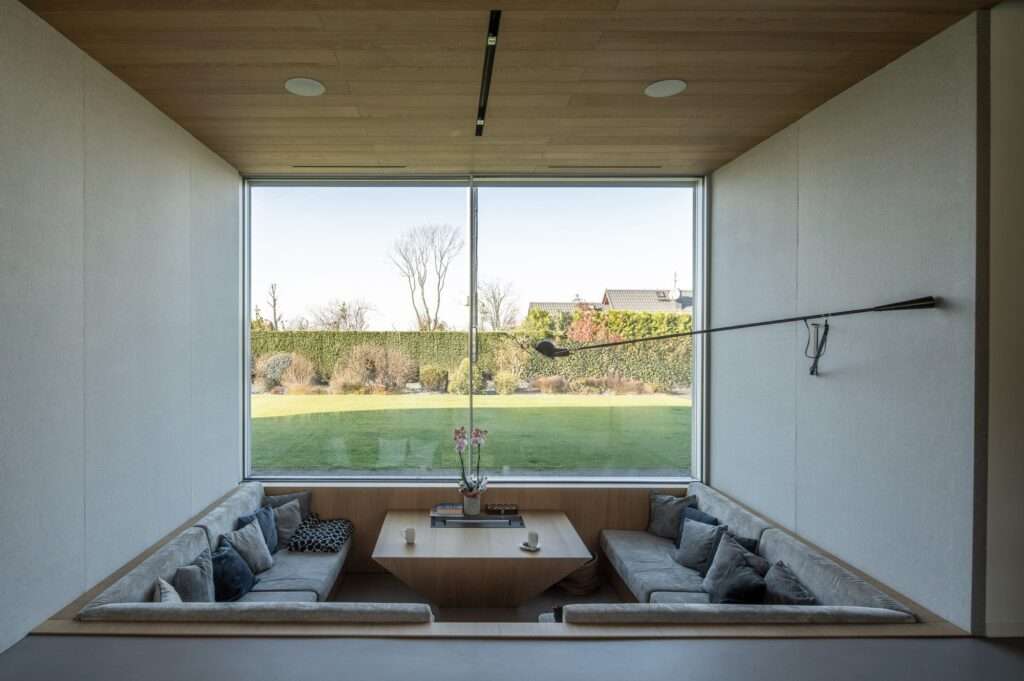 Interior shot of a cozy sunken conversation pit with grey plush sofas facing a massive panoramic window overlooking a green lawn.