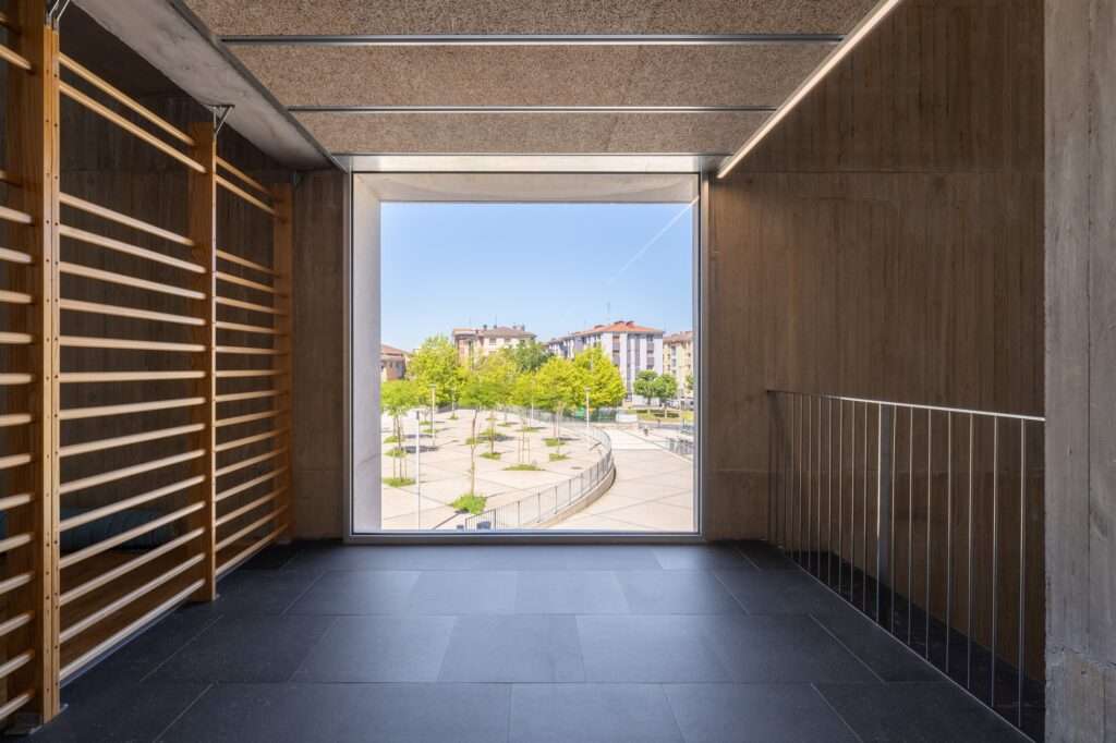 Interior view from a gym floor with wooden stall bars, looking through a large square window out onto the sunny public entry plaza and surrounding green trees.