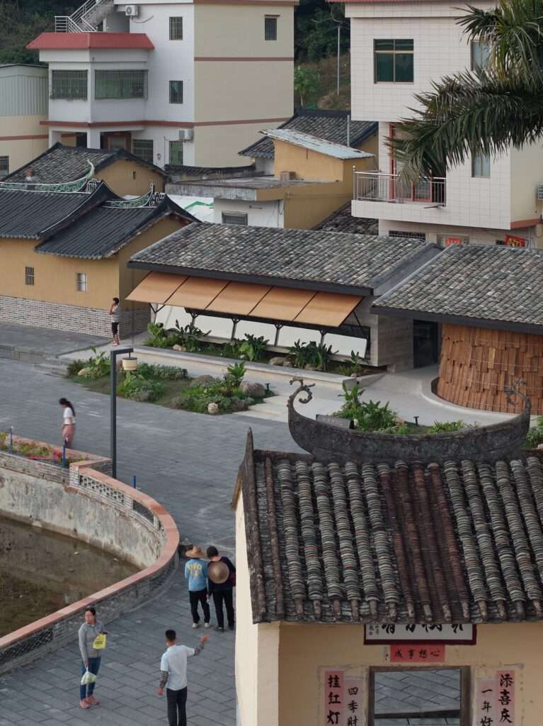 Elevated view overlooking the old village square and a traditional temple roof, with the newly renovated teahouse and tavern structures visible behind it.