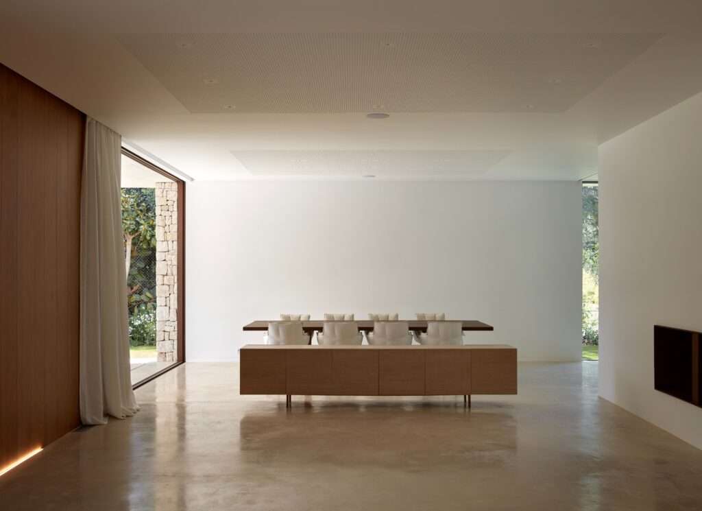 Spacious minimalist dining area with a long dark wooden table and a low wooden sideboard against a clean white wall.