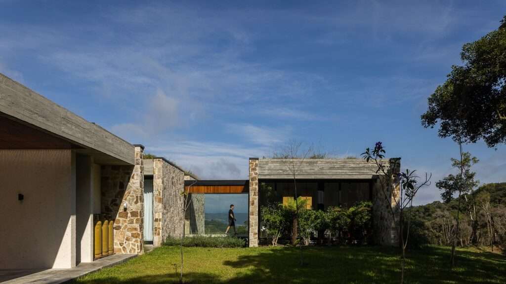 A glass-walled corridor link connecting two stone pavilions at Casa Inter Pires with a view of a person walking across.