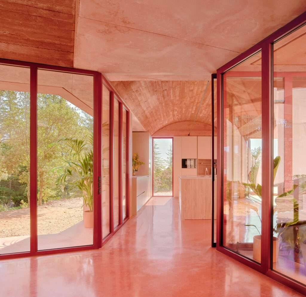 Glazed architectural corridor in Casa Sota la Mola with red frames, connecting different fragmented concrete volumes with views of the natural landscape.