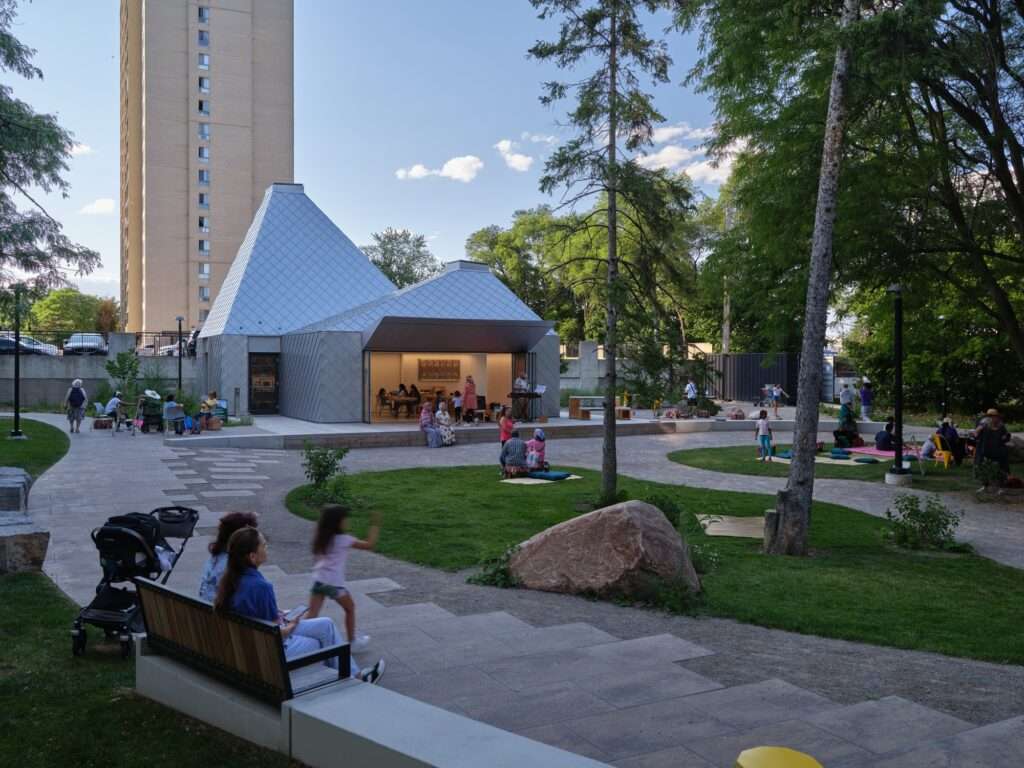 Landscaped public park with winding stone paths, green lawns, seating benches, and the modern aluminum pavilion set against residential high-rises.