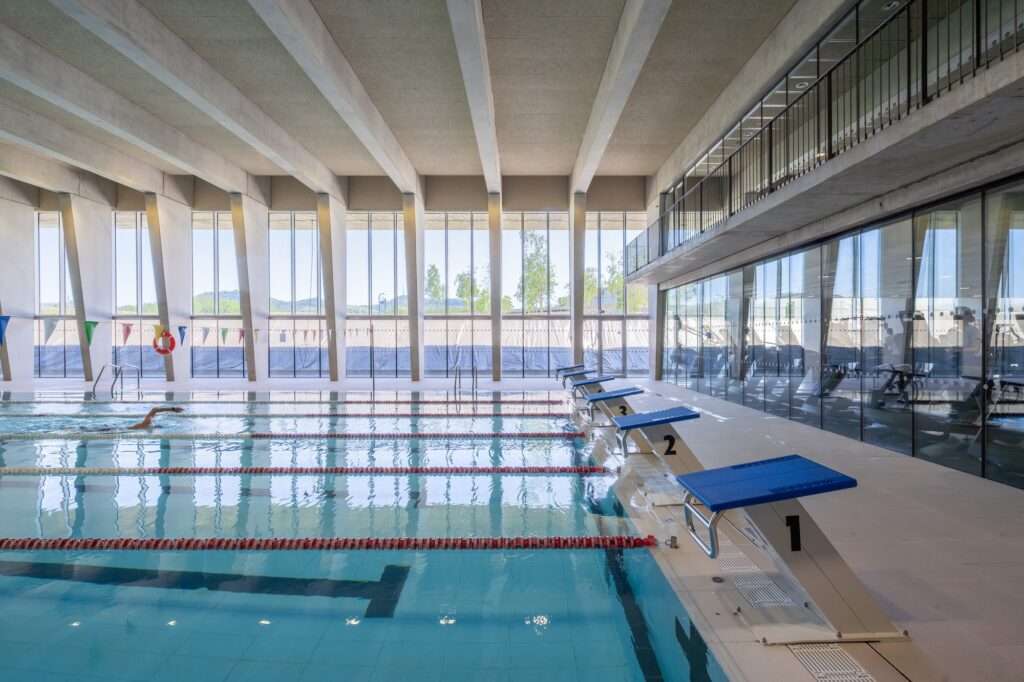 Indoor swimming pool area with blue starting blocks, concrete structural pillars, large south-facing windows, and a glass-walled gym overhead.