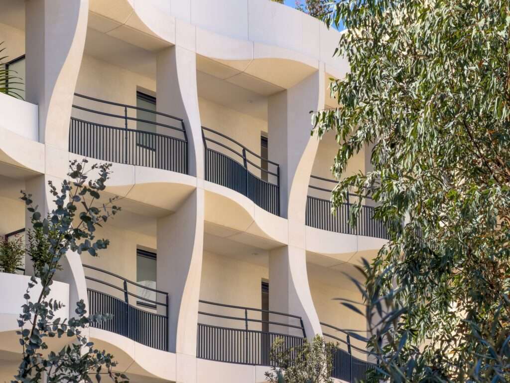 Close-up detail of white convex balconies and black railings at Secret Gardens, framed by the leaves of a Mediterranean tree.