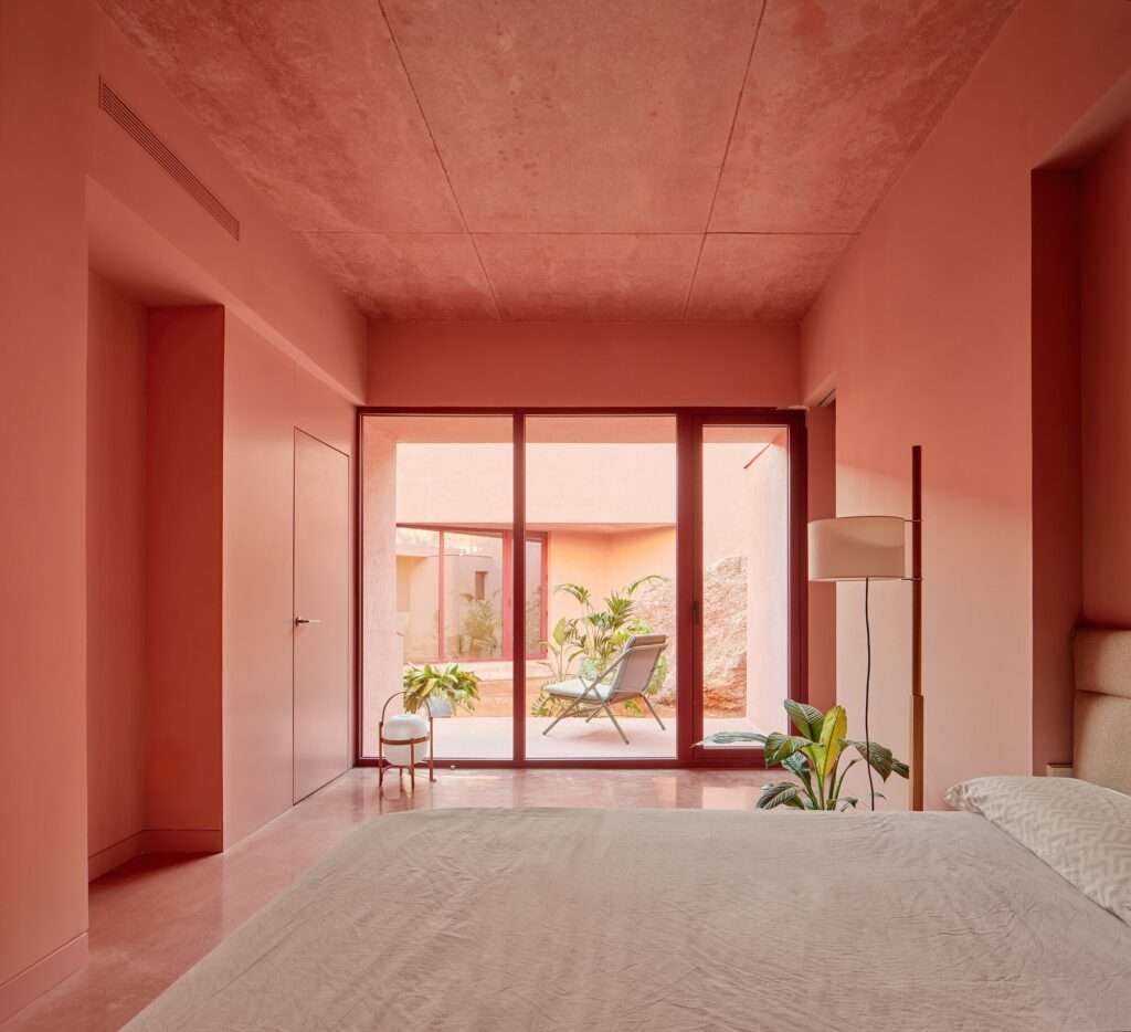 Minimalist master bedroom with red concrete walls and ceiling, looking through glass doors into a private central courtyard with outdoor seating.