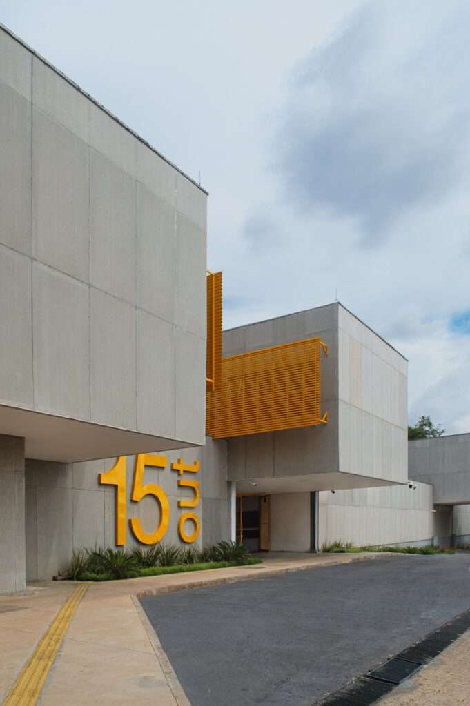 Angular low-angle view of the concrete school building showing modular cantilevered volumes, a bright yellow sign, and tactile paving on the sidewalk.