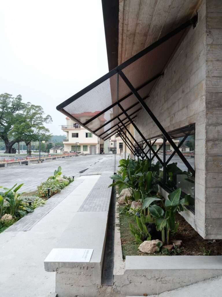 Exterior perspective along the facade of the Cave Teahouse, showing heavy steel-framed mesh awnings, planter boxes, and a concrete bench.