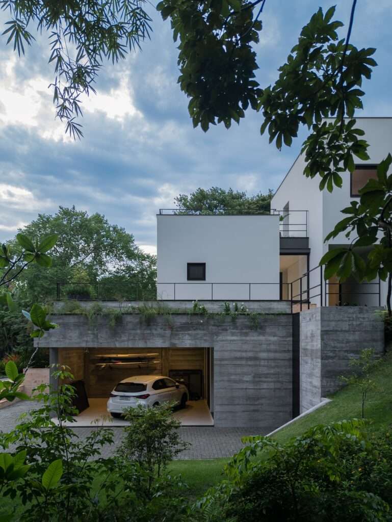 Evening view of a board-formed concrete garage integrated into the hill slope with a white car parked inside.