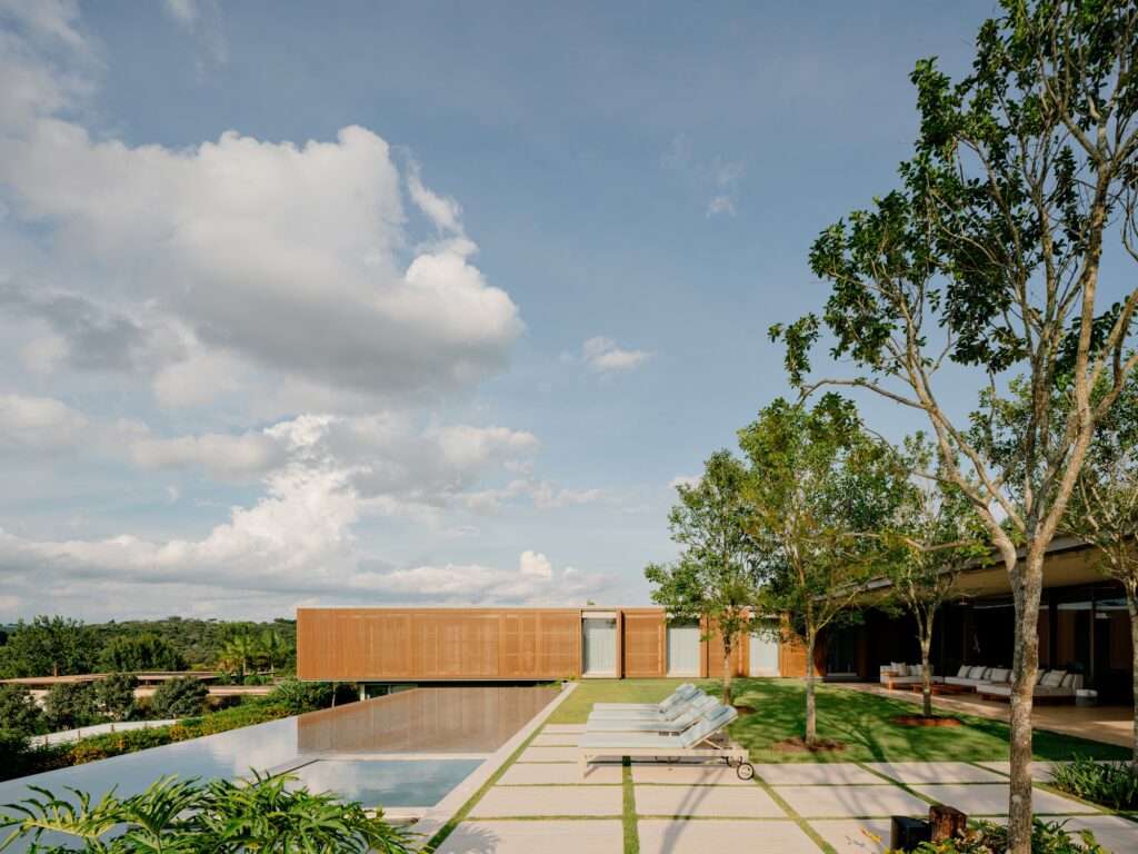 Panoramic view of Casa Escultura's L-shaped layout overlooking a flat lawn, infinity swimming pool, and lounge chairs against a blue sky.