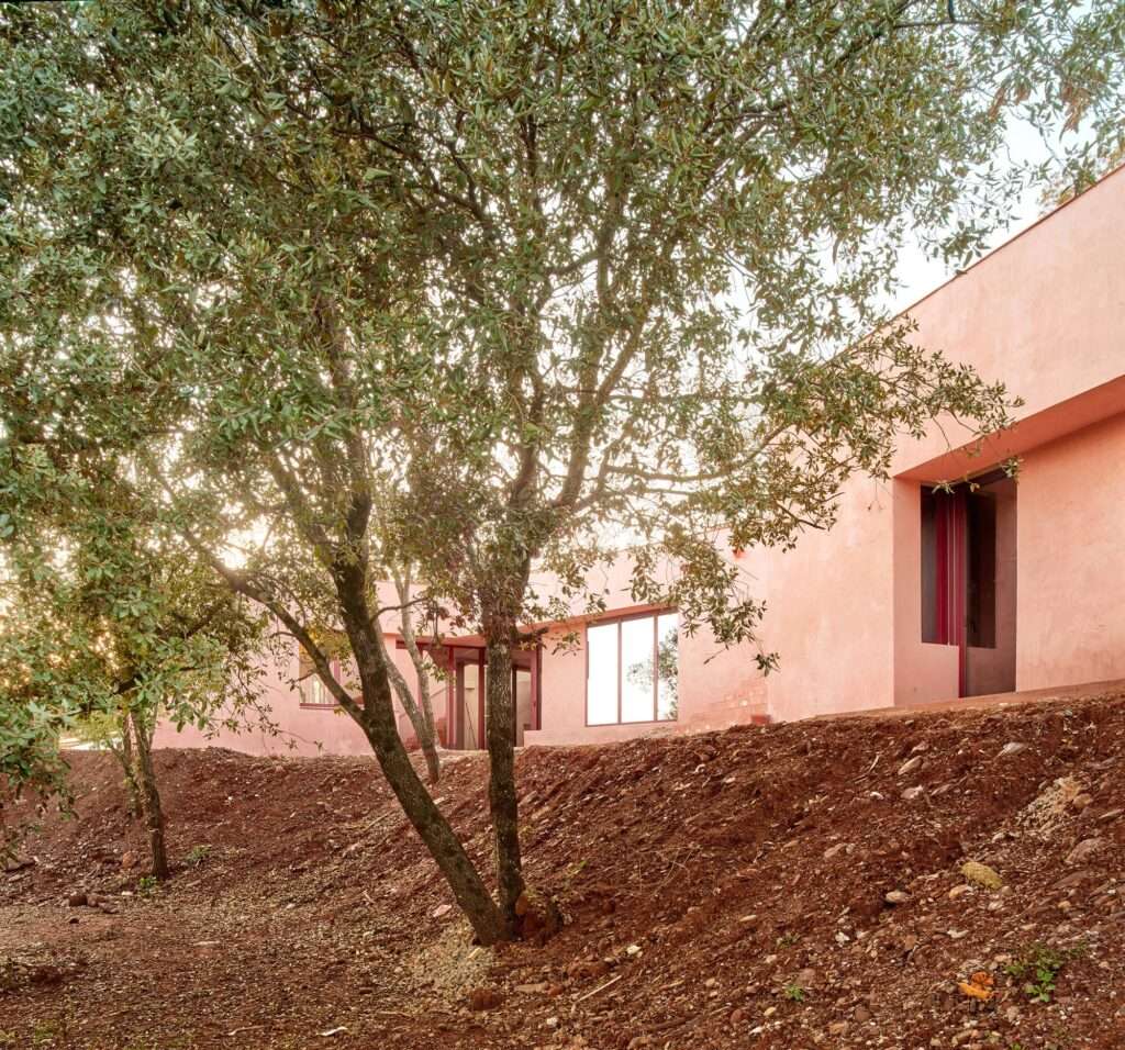 Red earth-toned concrete facade of Casa Sota la Mola situated on a steep sloped red dirt hillside with native trees.