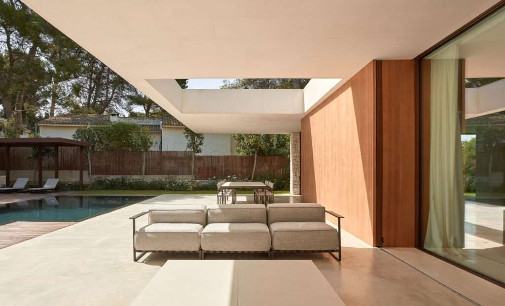 Covered outdoor patio with modern gray sofas and a dining table, protected by a white concrete cantilever with a rectangular opening.