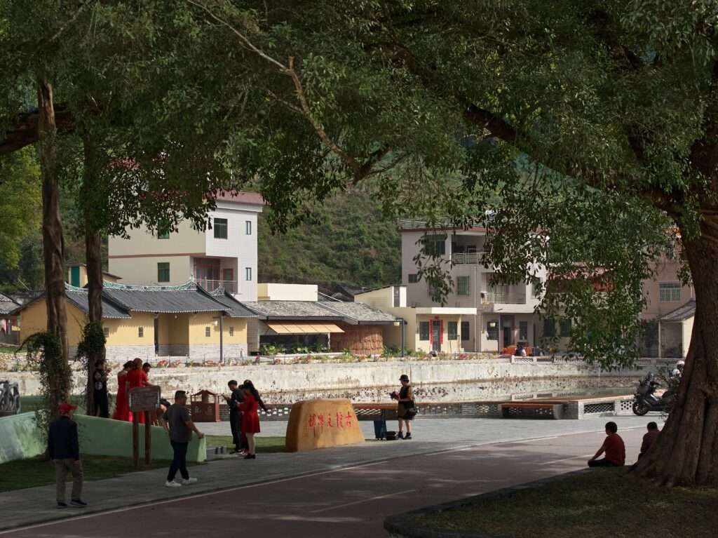 Distant landscape view of the village square across a pathway, framed by large ancient banyan trees with visitors walking around.