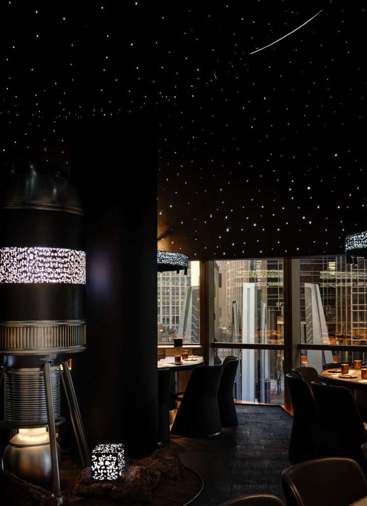 Restaurant interior view with an industrial metallic pod sculpture on a rocky base next to dining tables overlooking large floor-to-ceiling glass windows with city views.