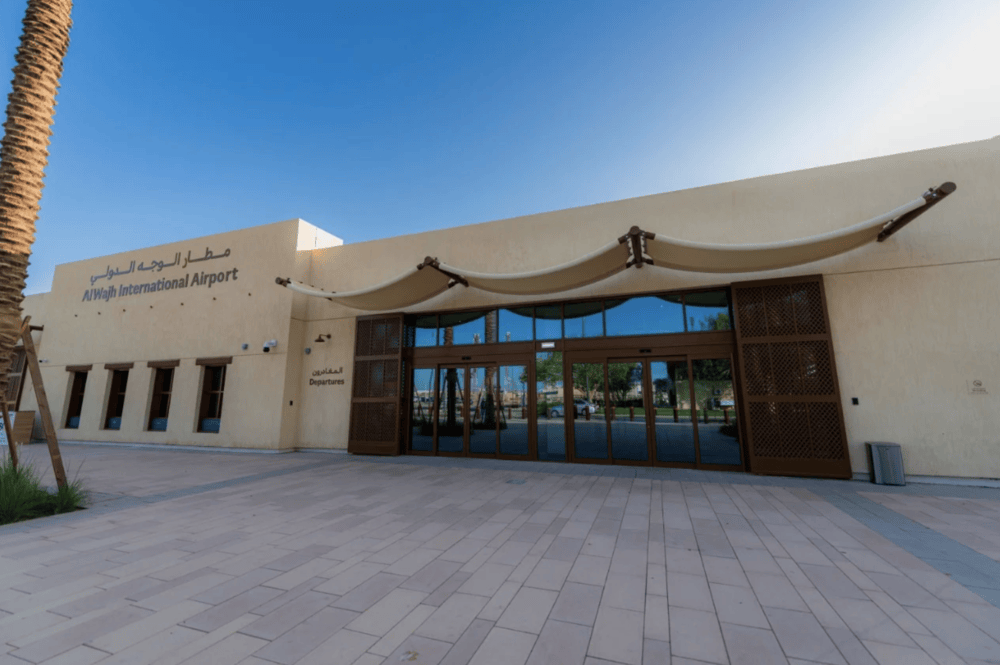 Exterior view of the AlWajh International Airport departures terminal with a large canopy and glazed enclosure under a clear blue sky.