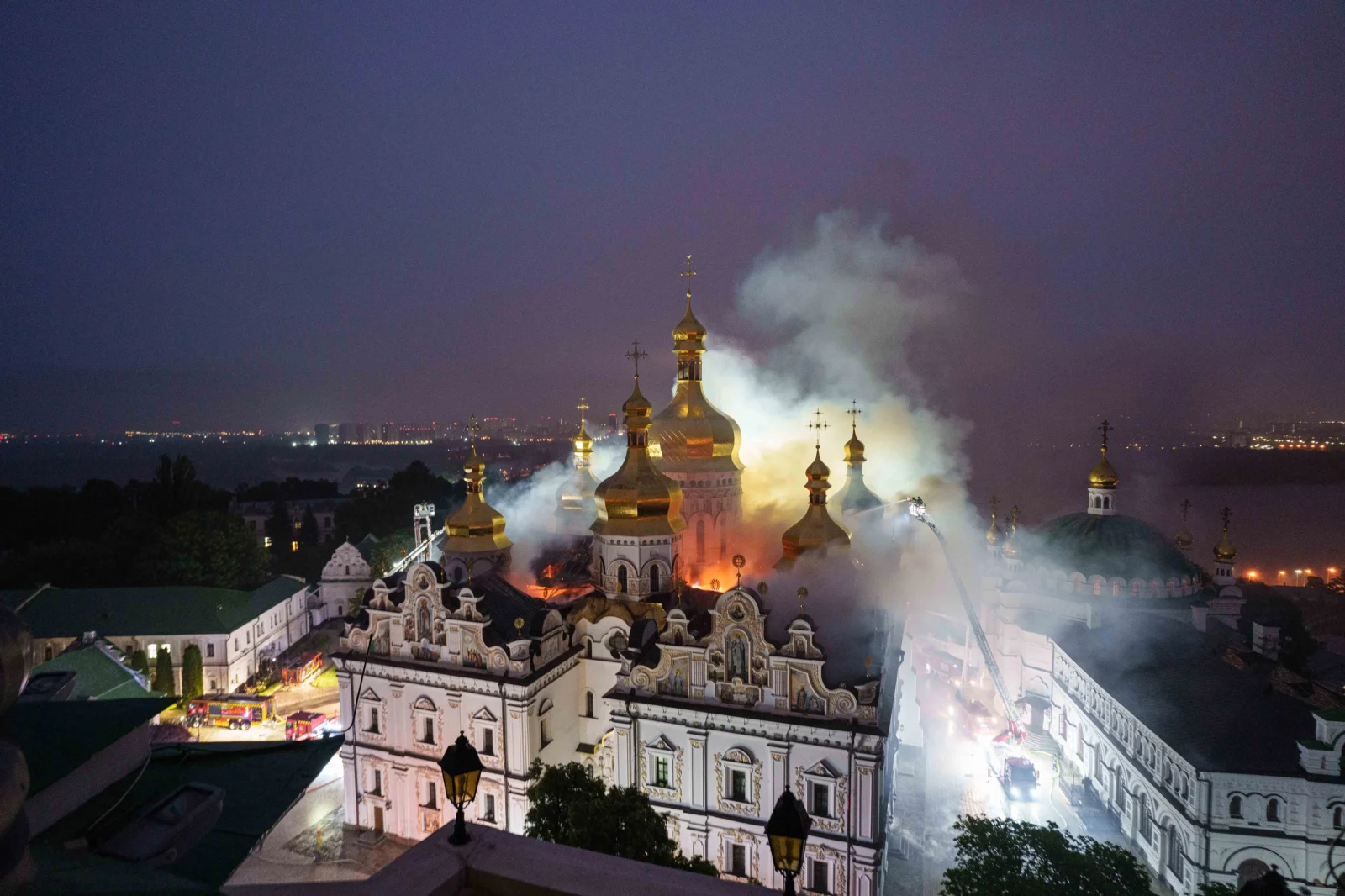Nighttime exterior view of the Dormition Cathedral at Kyiv Pechersk Lavra with smoke and fire rising from the roof structure.