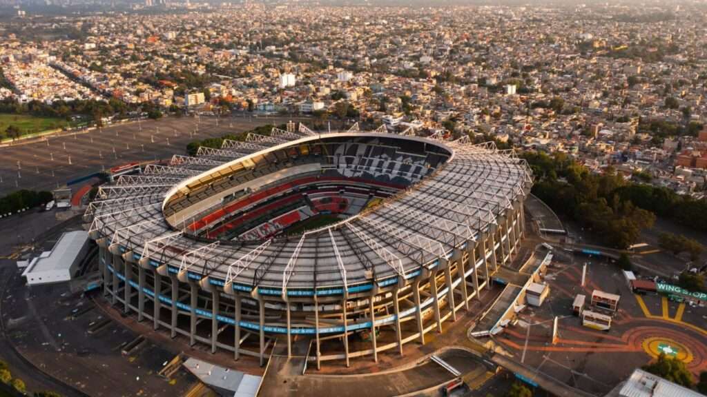 An aerial view showing the concrete massing and radial canopy structure of Azteca Stadium in Mexico City.