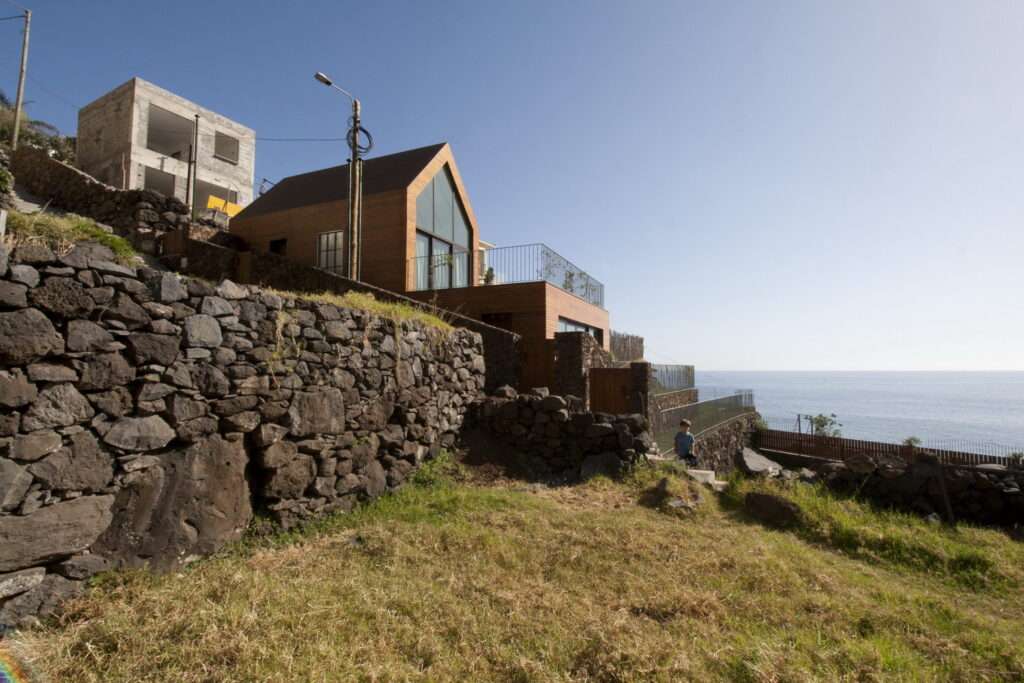 A high-angle exterior view looking across the wooden gabled roof of the upper house at Jardim do Mar. Solar panels are mounted on the lower section, and the house has views over the vast Atlantic Ocean and the vertical cliffs of the Madeira coastline.