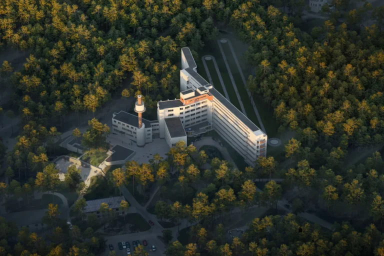 Aerial view of a white modernist building complex nestled within a dense forest of evergreen trees.