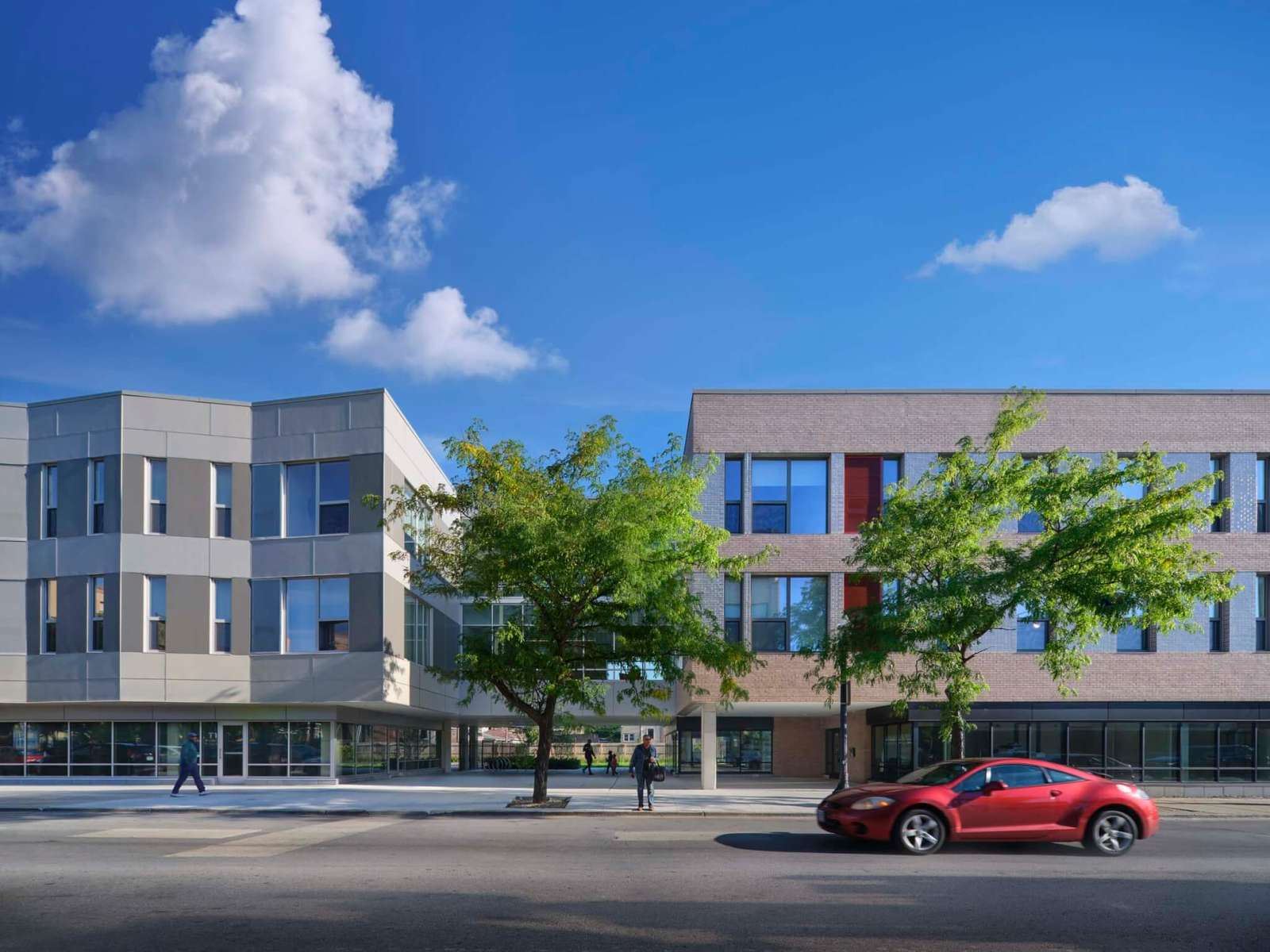 Two residential blocks with brick and undulating panel facades stand along a city street under a blue sky.