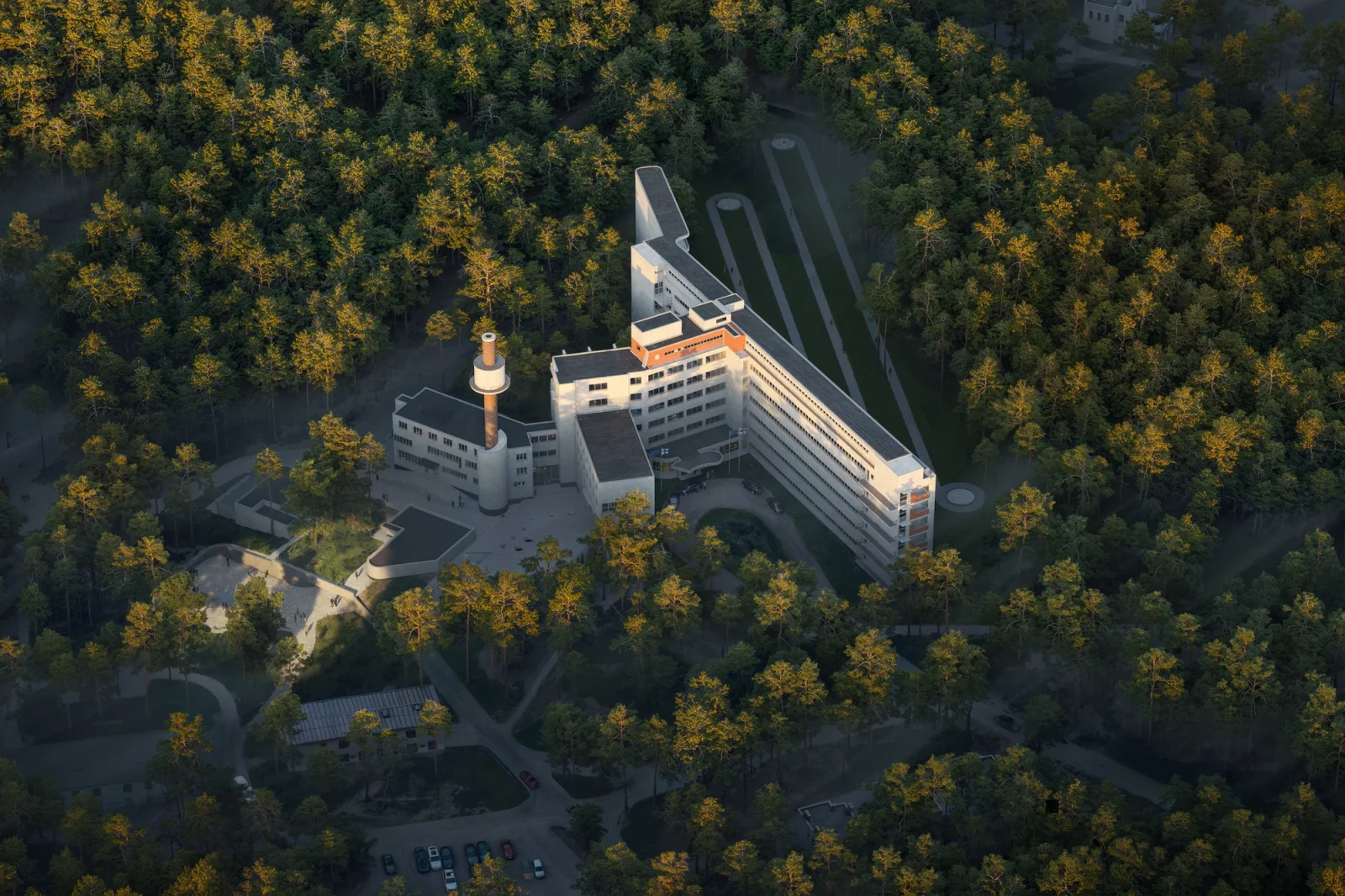 Aerial view of a white modernist building complex nestled within a dense forest of evergreen trees.