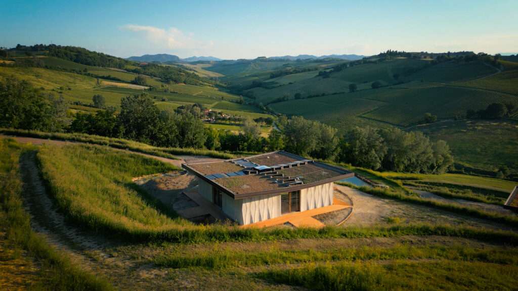 Aerial view of a single-story 3D-printed building with a green roof, situated on a grassy hillside.