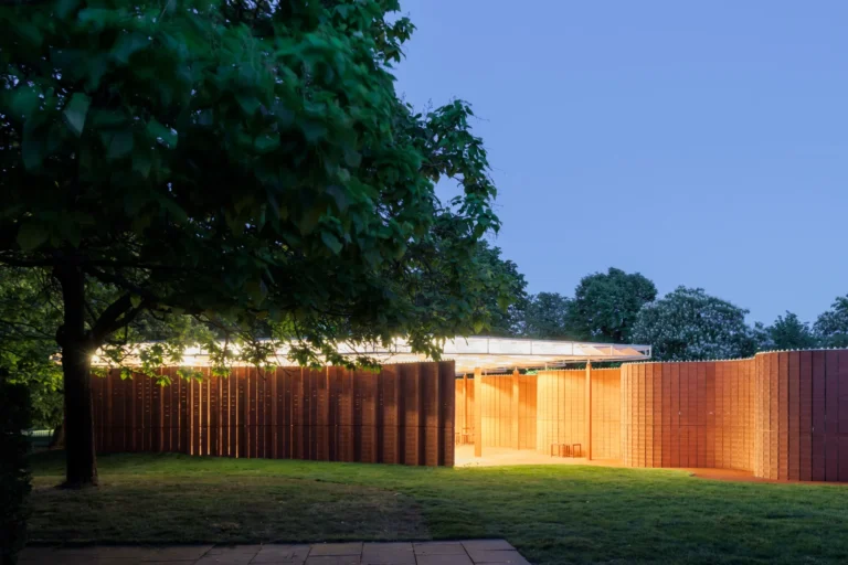 Night view of a curved brick pavilion illuminated from within, standing on a lawn next to a tree.