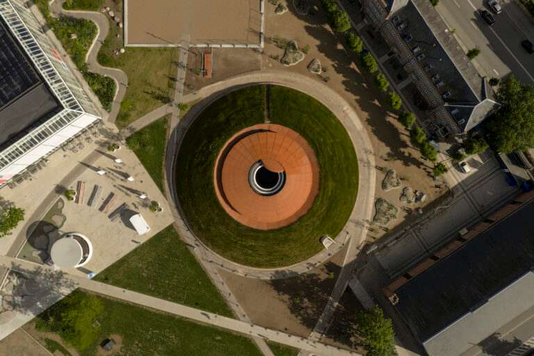 An aerial bird's-eye view of a circular mounded grass landscape containing a central copper-toned dome structure with a round opening.