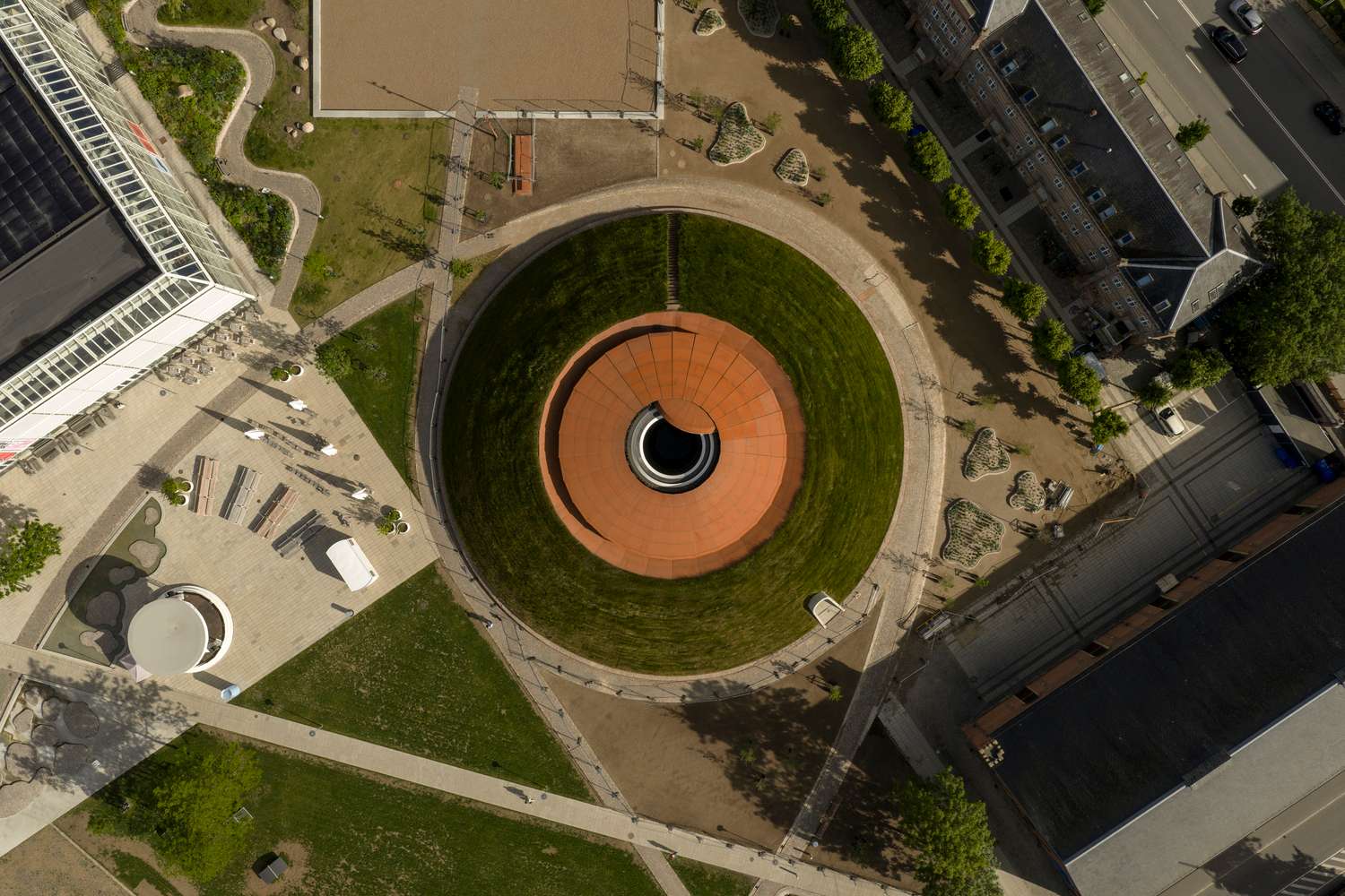 An aerial bird's-eye view of a circular mounded grass landscape containing a central copper-toned dome structure with a round opening.