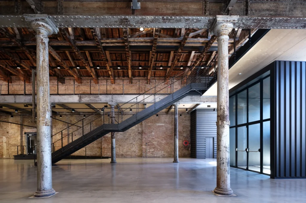 An expansive industrial interior showing historic columns, a steel staircase, and a modern glazed enclosure.