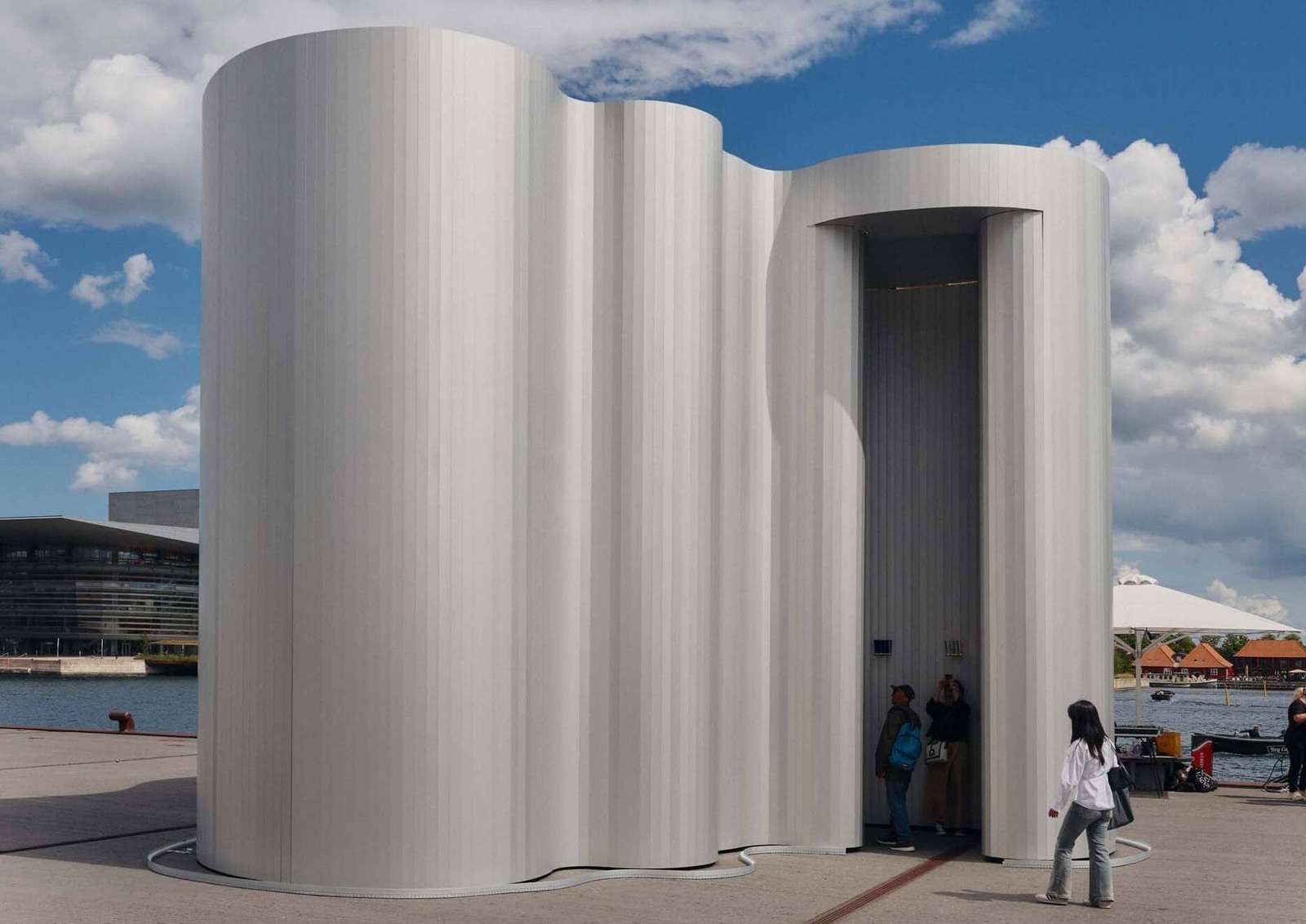 An exterior view of a tall, undulating aluminum pavilion situated on a waterfront plaza with visitors entering the threshold.