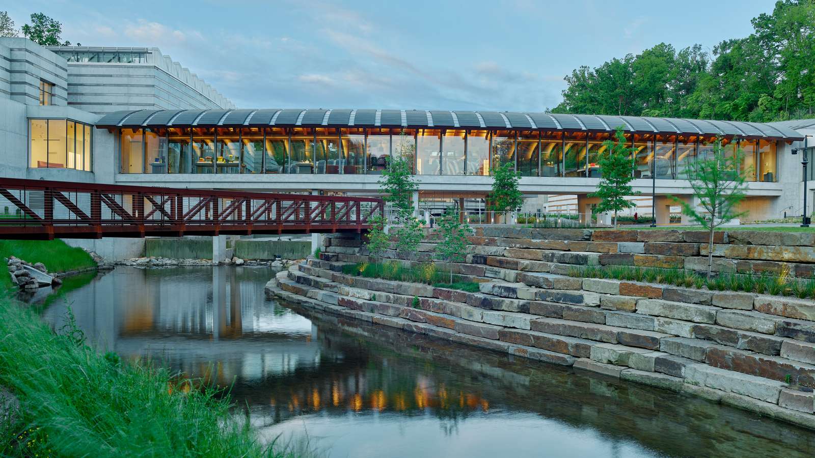 A glazed bridge gallery with a curved timber roof spans over a stream and tiered stone retaining walls.