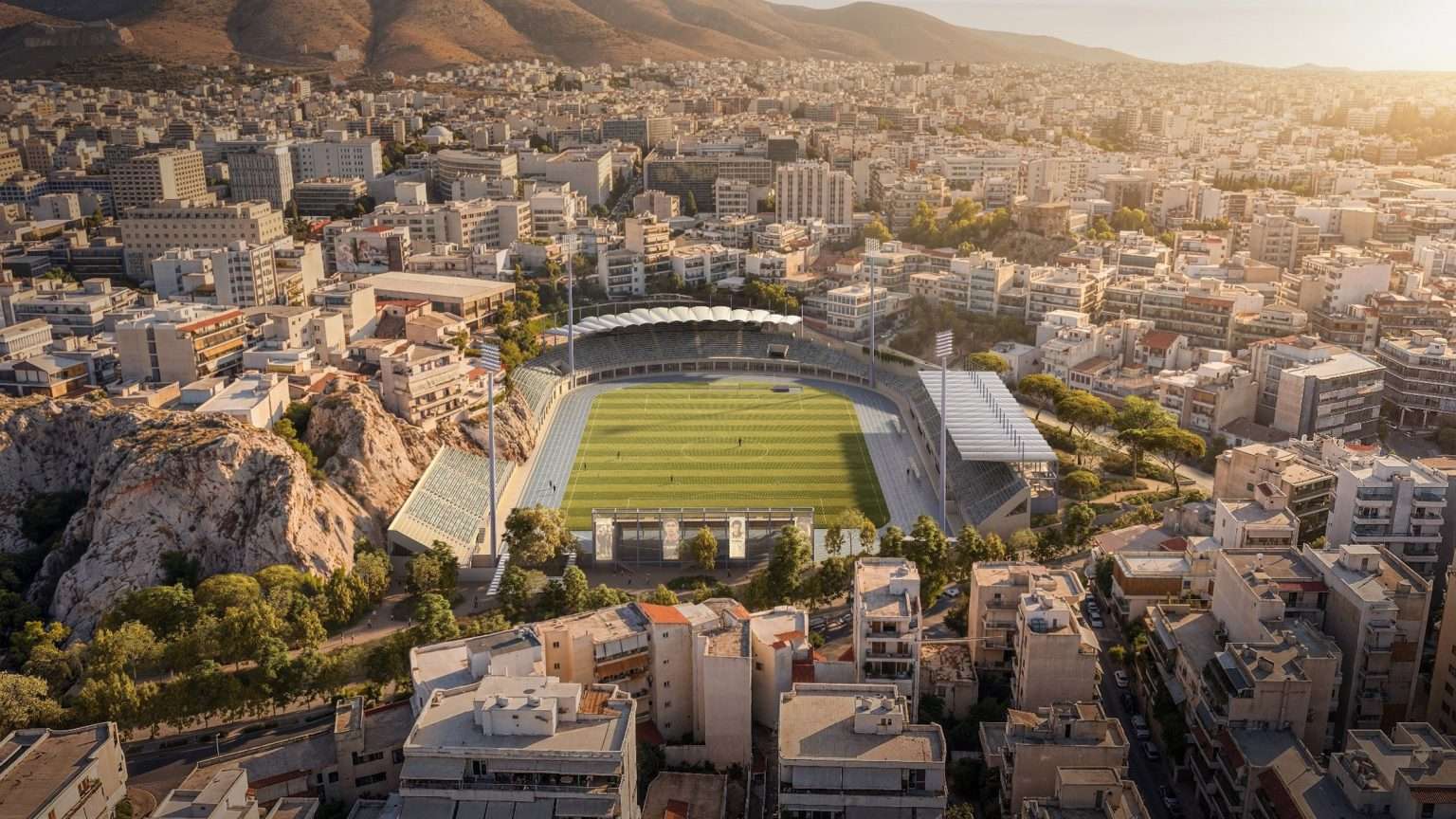Aerial view of an urban sports stadium surrounded by dense city blocks and a rocky outcrop.