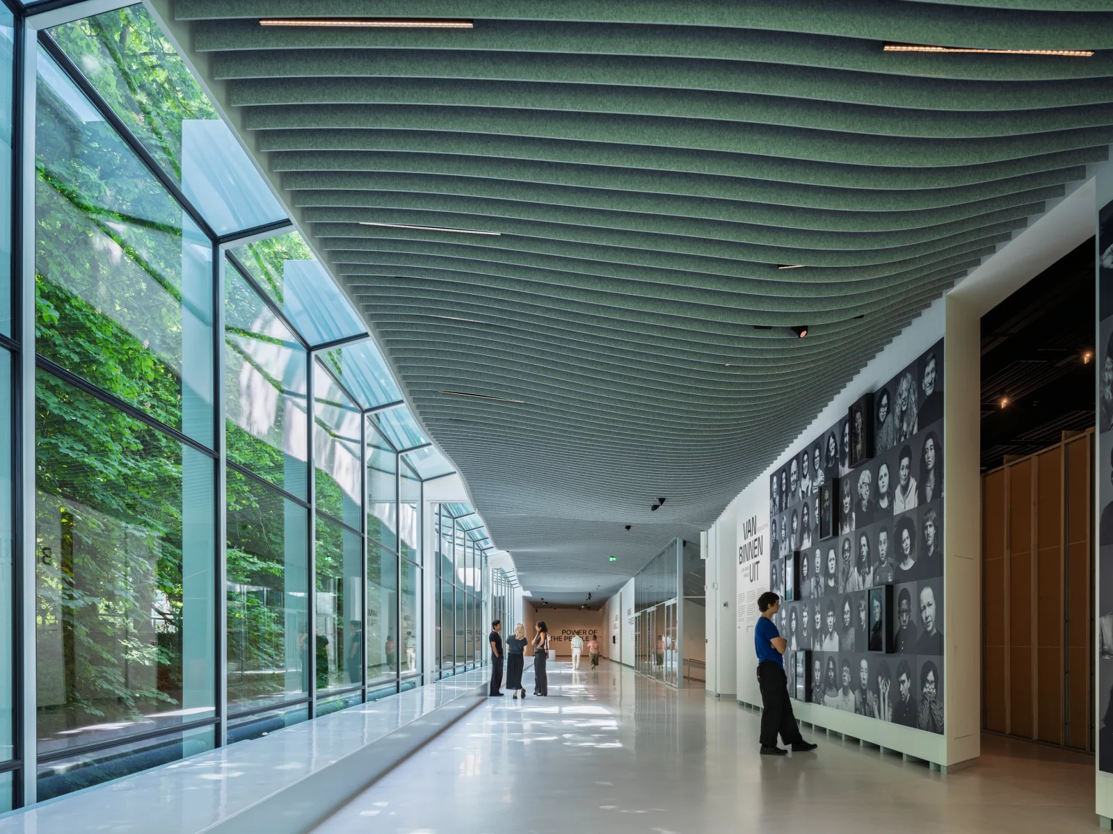 Interior view of a long museum hallway with an undulating acoustic ceiling and a glazed facade showing trees.